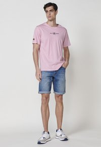 Rozā kokvilnas T-kreklu ar logo uz priekšpuses, pārī ar sarullētiem džinsu šortiem. Modelis valkā baltas un pelēkas sporta apavus. Vienkāršs dizains.