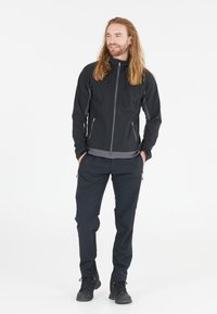 Chaqueta softshell negra con detalles en gris, con cremallera completa y capucha ajustable, combinada con pantalones negros y botas de montaña oscuras.