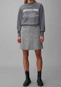 Sweatshirt gris avec texte blanc, associé à une jupe en laine grise avec une poche latérale et des bottines noires. Texture de tissu lisse.