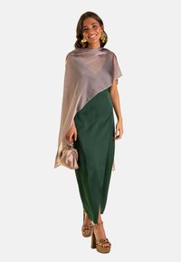 Vestido verde ajustado con una abertura lateral, acompañado de un chal transparente color topo. Sandalias de plataforma marrones con tiras y un bolso metálico completan el look.