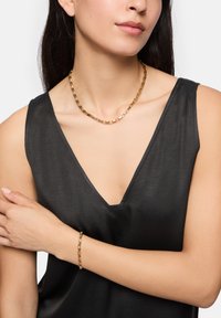 Gouden ketting met rechthoekige schakels, gedragen over een zwart mouwloos topje, met een gladde textuur en glanzende afwerking.