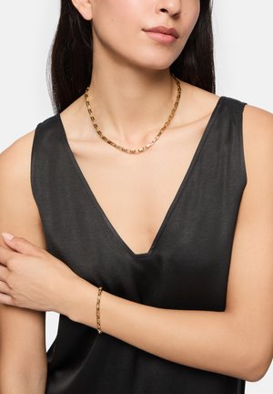 Gouden ketting met rechthoekige schakels, gedragen over een zwart mouwloos topje, met een gladde textuur en glanzende afwerking.