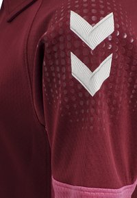 Bordeaux sportströja med texturerad tyg och cirkulära mönster, vita chevron-accenter och en rosa kant vid fållen.