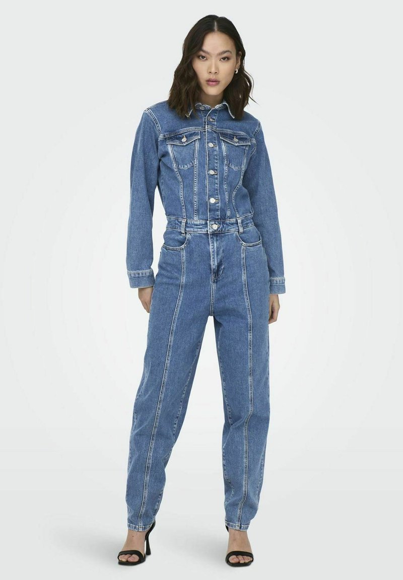 Femme debout portant une veste en denim bleue boutonnée et un pantalon taille haute assorti en denim, avec des sandales noires à talons hauts, sur fond blanc.