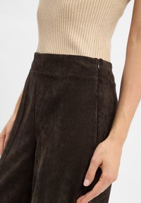 Un pantalon en velours côtelé marron avec des côtes verticales, une taille lisse et une fermeture éclair sur le côté. Le tissu a une apparence douce et texturée.