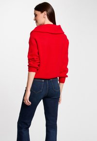 Rode gebreide trui met een brede kraag, gecombineerd met donkerblauwe slim-fit jeans. De trui heeft een gestructureerde stof en lange mouwen.