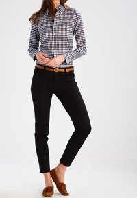 Camicia a quadretti bianco e nero con bottoni, pantaloni neri slim, cintura marrone e mocassini marroni. Il modello è in piedi con le mani sui fianchi.