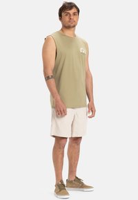 Olīvu zaļš bez piedurknēm krekls ar logotipu, apvienots ar krēmkrāsas šortiem un brūnām zamšādām kurpēm ar baltām zolēm. Viegla, ikdienišķa apģērba komplekts.