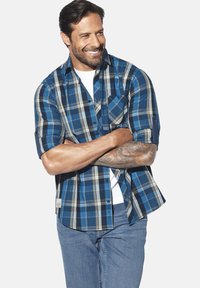 Mann mit tätowiertem Unterarm, der ein blaues karierter, hochgekrempeltes Hemd über einem weißen T-Shirt und blauen Jeans trägt, lächelt mit verschränkten Armen.