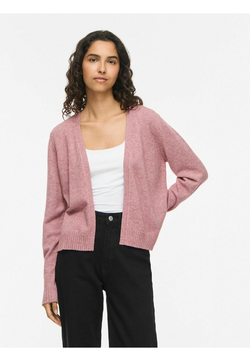 Femme aux cheveux bouclés et foncés, portant un cardigan rose clair, un top blanc et un pantalon noir, debout avec une main sur la hanche.