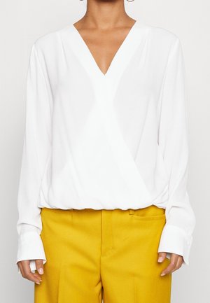 Vrouw draagt een witte blouse met lange mouwen en V-hals, ingestopt in hoog getailleerde mosterdgele broek, staand tegen een effen achtergrond.