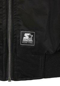 Veste noire à l'avant avec fermeture éclair argentée, poignet côtelé et patch logo en tissu rectangulaire Starter Black Label près de la fermeture éclair.