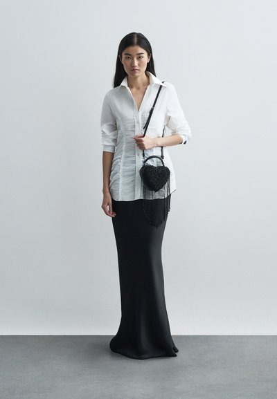 Γυναίκα που φοράει λευκό ριχτό πουκάμισο με κουμπιά και μακριές μαύρες φούστες, κρατώντας μια μαύρη τσάντα σε σχήμα καρδιάς με φούντες, μπροστά σε απλό φόντο.