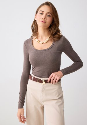 Kobieta o falistych brązowych włosach, ubrana w dopasowaną brązową bluzkę z długim rękawem, kremowe spodnie z brązowym paskiem oraz masywny naszyjnik z muszli, na jednolitym tle.