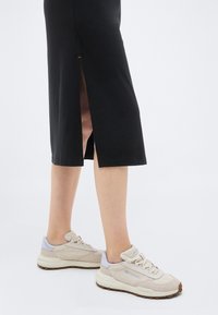 Κάτω άκρα ενός ατόμου που φοράει μπεζ αθλητικά παπούτσια και μαύρη φούστα με πλάγια σχισμή, στέκεται σε λευκό φόντο.