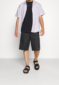 Camicia a maniche corte button-up di un leggero colore viola sopra una t-shirt nera, abbinata a pantaloni corti scuri e sandali neri con cinturini. Design e vestibilità semplici.