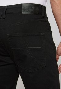 Jeans neri con una texture di tessuto liscia, dotati di una tasca posteriore con cuciture a zigzag e una patch in pelle del marchio sulla cintura.
