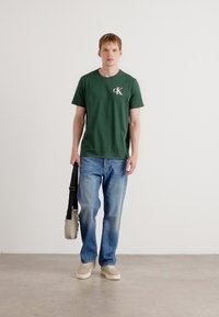 Grøn bomulds t-shirt med et hvidt logo, blå denim jeans, beige sneakers og en cremet skuldertaske, stående mod en ensfarvet baggrund.