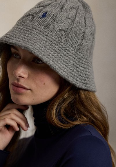 Šedý pletený klobouk typu bucket s texturovaným vzorem, zdobený modrým logem. Měkký materiál připomínající vlnu s širokou, dolů směřující krempou.