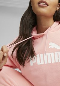 Roze hoodie met wit Puma-logo en tekst. Heeft een trekkoord en een zachte textuur. Hand van het model houdt het trekkoord vast, met lange, bruine haren zichtbaar.