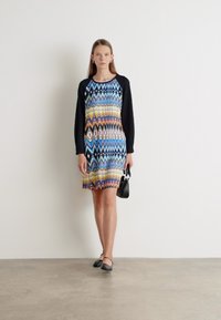 Vestido colorido con un patrón en zigzag en tonos azules, amarillos y negros, con largas mangas negras, complementado con un pequeño bolso negro.