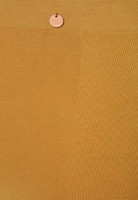 Texturierter senfgelber Stoff mit gerippten Muster und glatter Oberfläche. Ein runder Aufnäher mit dem Markennamen ist oben angebracht.