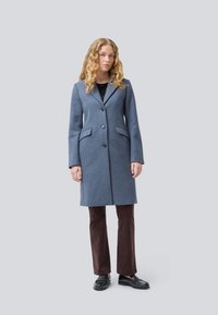 Wool blend coat in soft blue, featuring a notched collar, two buttons, and side pockets. Paired with brown corduroy pants and black shoes.
