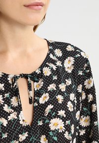 Blouse noire avec motif floral blanc et accents verts. Présente un décolleté en cœur avec un lien et un tissu doux et léger.