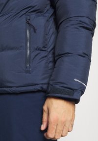 Marineblaue Daunenjacke mit gesteppte Abschnitten, Reißverschluss-Brusttasche und Klettverschluss an den Ärmeln. Mit "OMNI-HEAT"-Branding auf dem Ärmel.