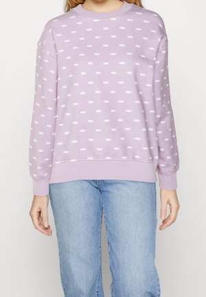 Osoba ubrana w lawendową bluzę z białymi prostokątnymi wzorami oraz jasnoniebieskie dżinsy, stojąca na jednolitym tle.