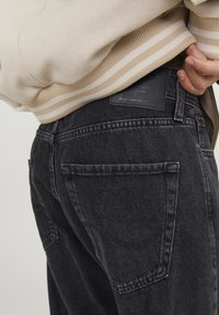 Cilvēks valkā melnus džinsus un krēmīgi baltu svītrainu džemperi, ar vienu roku ieliktu aiz muguras pie vidukļa.