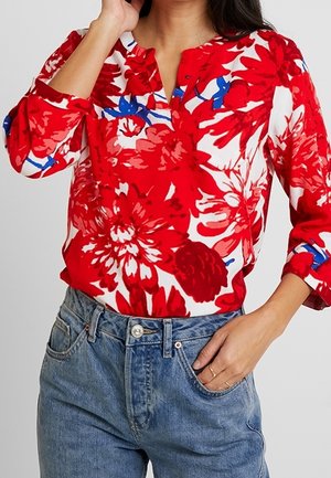 Femme portant un chemisier à fleurs rouge, blanc et bleu avec des manches 3/4, rentré dans un jean bleu taille haute, main dans la poche.