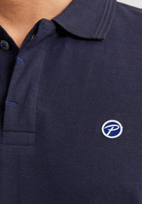 Námornická polo košile s texturovanou tkaninou, límcem na dva knoflíky a malým kulatým logem v modré a bílé barvě na levém prsu.