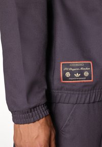 Αθλητικό μπουφάν FC Bayern München σε σκούρο μοβ ύφασμα, με ριμπ μανσέτες, τσέπες και έμβλημα με χρυσές λεπτομέρειες.