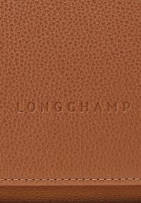 Pelle marrone testurizzata con la parola LONGCHAMP impressa al centro sopra una cucitura, in primo piano.