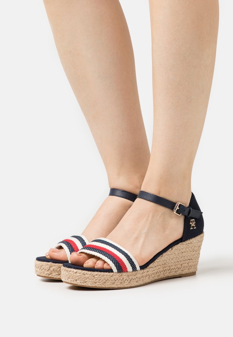 Pieds portant des sandales compensées bleu marine avec des brides en tissu rayé rouge, blanc et bleu marine et des fermetures à boucle à la cheville, posés sur une surface blanche.