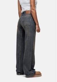 Jeans en denim foncé à jambes larges avec taille haute, dotés de poches arrière et d'un patch en cuir à la taille. Texture lisse, design décontracté.