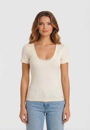 Donna con capelli castano chiaro che indossa una maglietta crema a maniche corte con scollatura rifinita in pizzo e jeans azzurri, in piedi davanti a uno sfondo semplice.