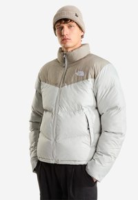 Млад мъж, носещ светлосиво и бежово пухено яке North Face и бежова плетена шапка, стои с ръце в джобовете.