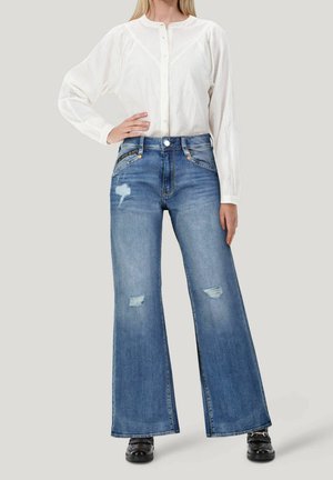 Vrouw die een wit langärmelig blouse met knopen draagt en een high-waisted wijde blauwe jeans met versleten plekken, poseert met een hand op haar heup.