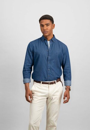 Camisa de mezclilla de botones en azul oscuro con mangas largas y puños con botones, combinada con pantalones rectos de color crema y un cinturón trenzado marrón.