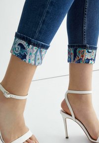 Piernas inferiores de una persona que lleva puestos unos pantalones vaqueros cortos azul con puños de paisley colorido y sandalias blancas de tacón alto con tiras al tobillo.