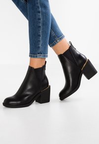 Botines de cuero negro con tacón cuadrado, que cuentan con paneles laterales elásticos y un detalle de acento en dorado. Textura suave y diseño de punta redonda.