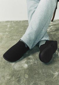 Juodi slip-on batai su žakardo tekstūra, guminiais padais, derinami su pilkais sportiniais kelniais, padėti ant tekstūruoto žalių kilimėlių.