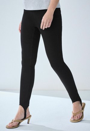 Femme portant un legging noir à étriers et des sandales à talons couleur beige marchant sur un fond clair.