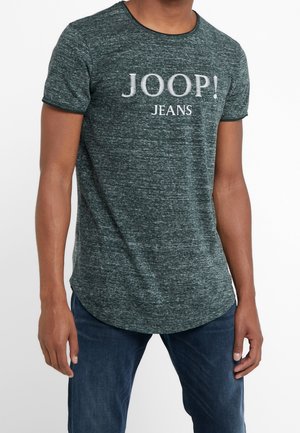 T-shirt vert foncé à manches courtes en tissu texturé, avec le texte blanc "JOOP! JEANS". L'ourlet courbé ajoute une forme distincte.