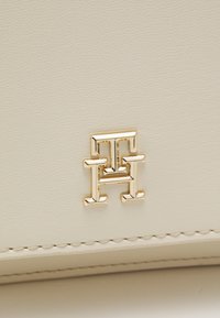 Borsa in pelle crema con superficie testurizzata, caratterizzata da un prominente emblema dorato e cuciture precise lungo i bordi.