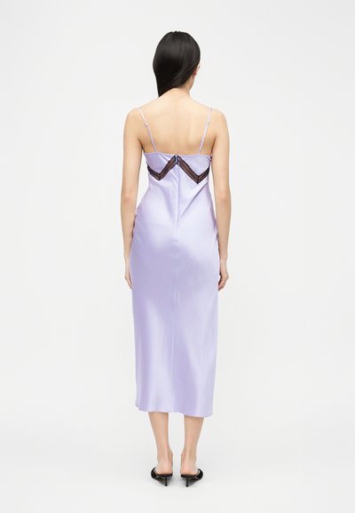 Γυναίκα που στέκεται με την πλάτη, φορώντας λιλά σατέν φόρεμα με λεπτές τιράντες και μαύρη δαντέλα στην πλάτη, συνδυασμένο με μαύρες γόβες.