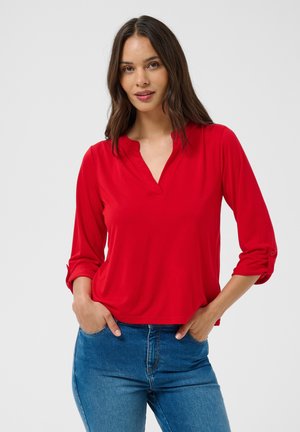 Jeune femme aux longs cheveux foncés portant un haut rouge à manches longues et un jean bleu, debout les mains dans les poches devant un fond clair.