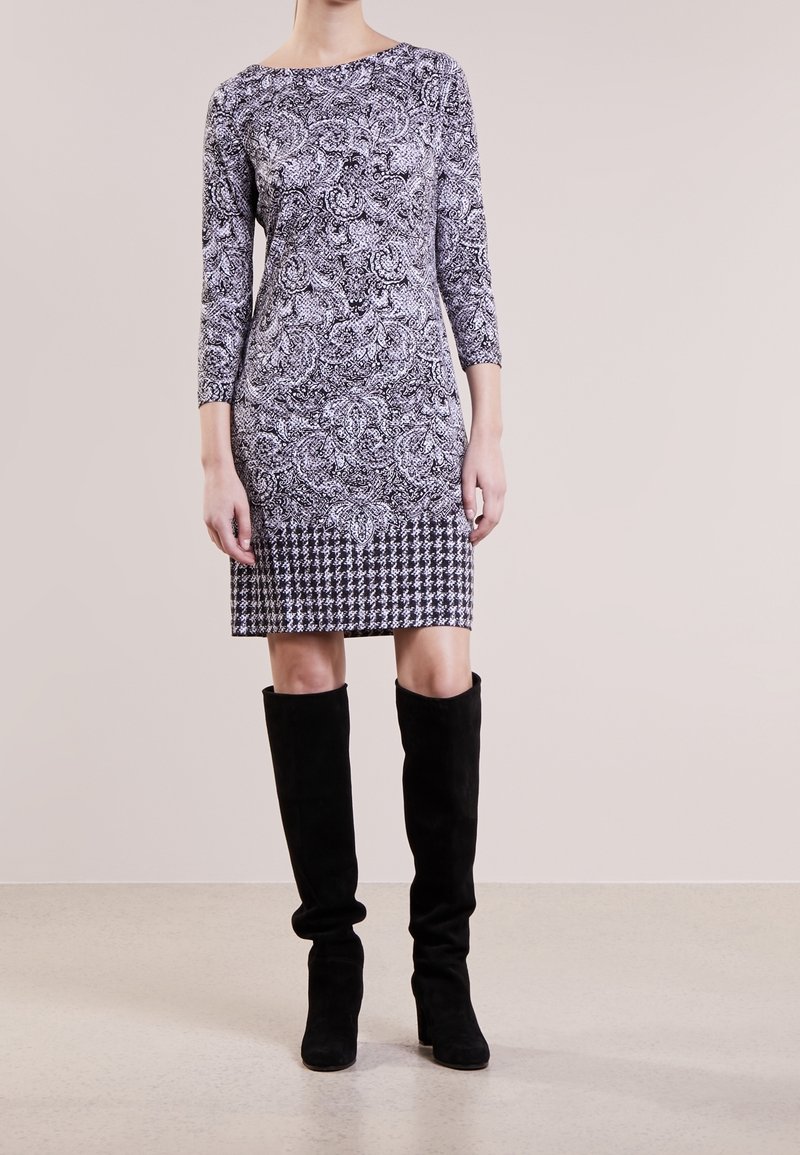 Femme portant une robe moulante longueur genou avec des motifs cachemire et pied-de-poule, ainsi que des bottes noires en daim montant jusqu'au genou, debout sur un sol uni.